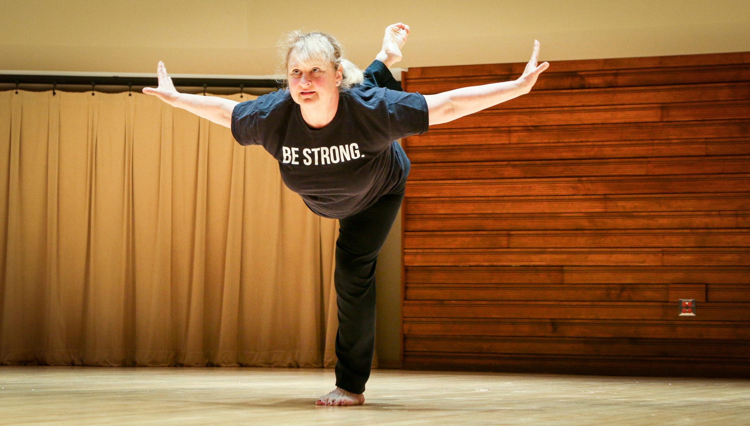 woman balancing on one leg in a flying pose, her tshirt reads, "Be Strong".