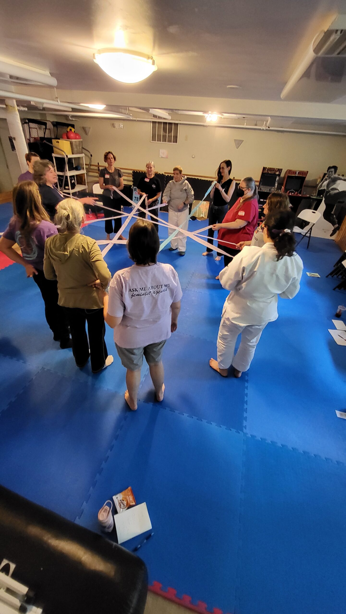 A group of class participants standing in a circle. They are  each holding one end of flat, belt-like bands, and a person across from them holds the other end. The bands all weave together within the circle.